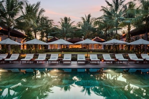 A serene resort setting features a series of lounge chairs lined in a row beside a reflective swimming pool. Large umbrellas provide shade, set against a backdrop of tall palm trees and a warm, glowing structure with expansive windows. The early evening sky casts a soft, calming glow over the entire scene.