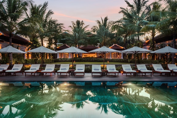 A serene resort setting features a series of lounge chairs lined in a row beside a reflective swimming pool. Large umbrellas provide shade, set against a backdrop of tall palm trees and a warm, glowing structure with expansive windows. The early evening sky casts a soft, calming glow over the entire scene.