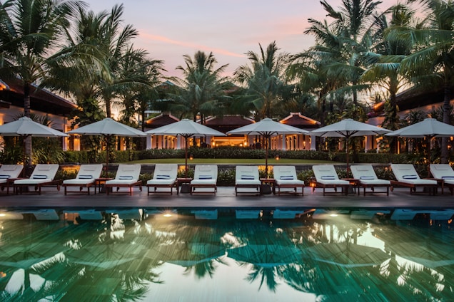 A serene resort setting features a series of lounge chairs lined in a row beside a reflective swimming pool. Large umbrellas provide shade, set against a backdrop of tall palm trees and a warm, glowing structure with expansive windows. The early evening sky casts a soft, calming glow over the entire scene.