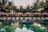 A serene resort setting features a series of lounge chairs lined in a row beside a reflective swimming pool. Large umbrellas provide shade, set against a backdrop of tall palm trees and a warm, glowing structure with expansive windows. The early evening sky casts a soft, calming glow over the entire scene.