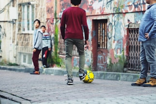 A group of children playing soccer in an urban street setting. The scene includes graffiti-covered walls and worn-out buildings, giving a rustic and lively atmosphere. The children appear relaxed and engaged, with one of them balancing a soccer ball at his feet.
