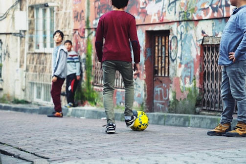 A group of children playing soccer in an urban street setting. The scene includes graffiti-covered walls and worn-out buildings, giving a rustic and lively atmosphere. The children appear relaxed and engaged, with one of them balancing a soccer ball at his feet.