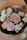 A bouquet of flowers featuring a variety of blooms, including pastel pink and white roses, a pink carnation, and white lisianthus. The flowers are accompanied by green leaves and are wrapped in brown paper.