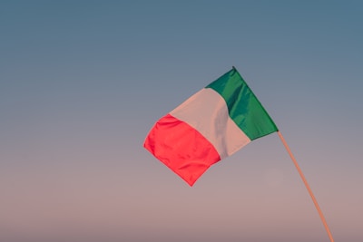 The Italian flag waving gently against a bright blue sky over farmland.