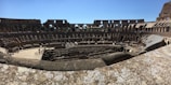 An ancient amphitheater with large open arches and a partially intact structure, revealing multiple levels of seating and the central arena floor. Several tourists are visible exploring the site under a clear blue sky.
