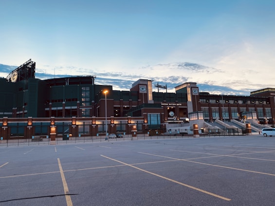 A large, iconic sports stadium with the Green Bay Packers logo prominently displayed, surrounded by an empty parking lot. The stadium is constructed with green and brown elements, and the sky above is partly cloudy with soft evening light.