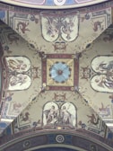 A richly decorated ceiling with intricate frescoes depicting mythological figures and ornate designs. The central motif features a starburst pattern surrounded by symmetrical, detailed imagery, including human figures, mythical creatures, and elaborate floral patterns. The artwork is set within a structured frame of geometric shapes and borders, showcasing Renaissance-style artistry.