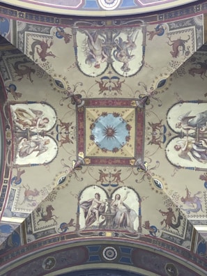 A richly decorated ceiling with intricate frescoes depicting mythological figures and ornate designs. The central motif features a starburst pattern surrounded by symmetrical, detailed imagery, including human figures, mythical creatures, and elaborate floral patterns. The artwork is set within a structured frame of geometric shapes and borders, showcasing Renaissance-style artistry.