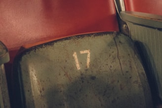 A close-up view of a vintage, worn-out stadium seat labeled with the number 17. The seat is made of metal and appears to have a greenish hue, showing signs of rust and wear. Adjacent to the seat is a red background that could be another seat or the wall of the stadium.