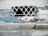 A weathered and deteriorating wall with peeling paint and a small, decorative metal grille. The paint is chipped, revealing layers of blue and white beneath. The wall appears old and in need of maintenance, with rough textures visible.