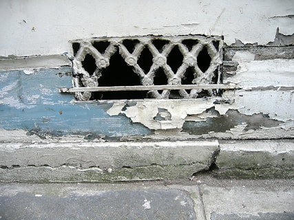 A weathered and deteriorating wall with peeling paint and a small, decorative metal grille. The paint is chipped, revealing layers of blue and white beneath. The wall appears old and in need of maintenance, with rough textures visible.