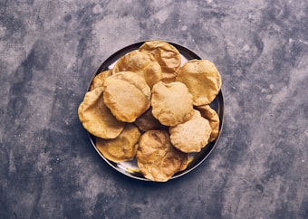A metal plate holds a stack of golden-brown fried flatbreads arranged neatly in a circular fashion. The background is a textured grey surface, providing contrast to the warm tones of the bread.