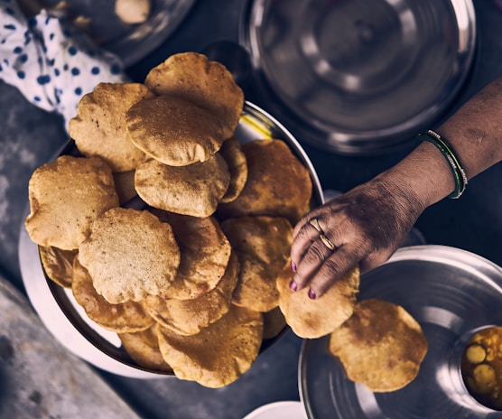A person with bangles on their wrist is picking up a fried bread from a stack of similar pieces in a large metal bowl. Various kitchen utensils and dishes are also visible in the background, including a cloth with blue dots.