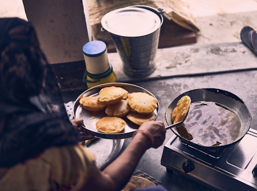 A person is frying round, golden-brown bread-like food in a pan filled with oil on a stove. Nearby, there is a metal bucket, a container with a blue lid, and a stack of fried bread placed on a round tray being held by the person.