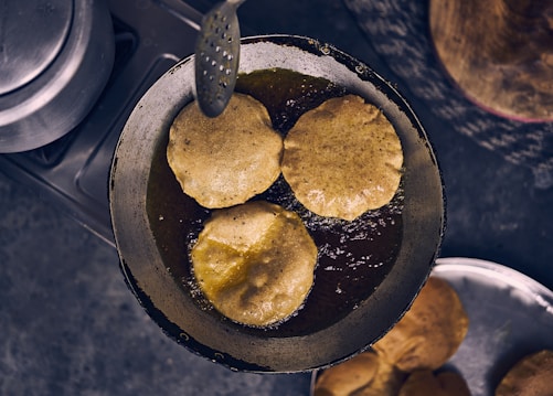 Three pieces of bread are being deep-fried in hot oil inside a pan on a stovetop. The bread appears to be puffed up, indicating they are puris or similar fried dough. Nearby, there is a slotted spoon for handling the food, and a metal lid and plate are visible in the background.