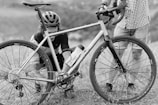 Two individuals are outdoors with a bicycle, one of whom is kneeling while working on the bike. The person kneeling is wearing a helmet and cycling gear while the other is standing with one hand on the handlebars, dressed in a checkered shirt and shorts.
