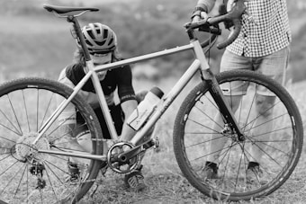 Two individuals are outdoors with a bicycle, one of whom is kneeling while working on the bike. The person kneeling is wearing a helmet and cycling gear while the other is standing with one hand on the handlebars, dressed in a checkered shirt and shorts.