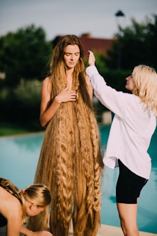 A person stands by a poolside with extremely long, flowing hair, while another person to the right appears to be styling or adjusting the hair. A third person is crouched nearby, possibly assisting in the styling process. The scene is set outdoors with trees and a building in the background, suggesting a serene and relaxed setting.