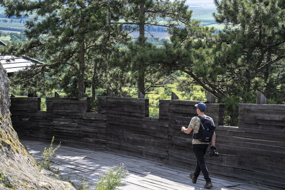 A person with a backpack and a camera is walking along a wooden observation deck surrounded by dense green trees. The scene depicts a natural setting with a wooden barrier that allows for a view of the surrounding landscape.