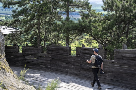 A person with a backpack and a camera is walking along a wooden observation deck surrounded by dense green trees. The scene depicts a natural setting with a wooden barrier that allows for a view of the surrounding landscape.
