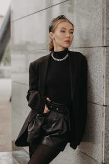 A stylish individual with a sleek hairstyle, wearing a black outfit with a chain necklace, leans confidently against a gray stone wall. The outfit includes a blazer, high-waisted shorts, and stockings.
