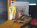 A playful kitten is lying on a wooden surface with its mouth open, appearing as if it's meowing or yawning. Next to the kitten is a yellow package, likely a cleaning product, and in the background, a cardboard box with blue text.