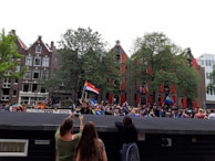A lively outdoor event with a crowd of people holding various colorful flags. The setting appears to be in a city with traditional brick buildings and trees in the background. Some participants are taking photos, and there is a festive atmosphere.