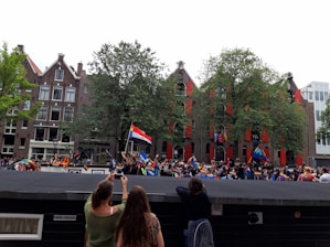 A lively outdoor event with a crowd of people holding various colorful flags. The setting appears to be in a city with traditional brick buildings and trees in the background. Some participants are taking photos, and there is a festive atmosphere.