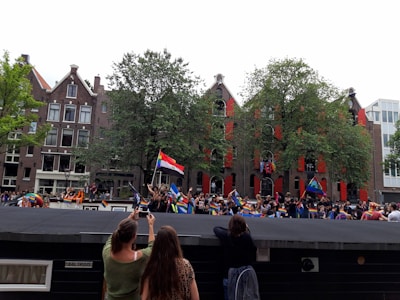 A lively outdoor event with a crowd of people holding various colorful flags. The setting appears to be in a city with traditional brick buildings and trees in the background. Some participants are taking photos, and there is a festive atmosphere.