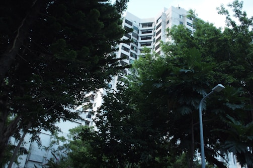 A tall, modern apartment building is partially obscured by lush green trees, creating a blend of urban and natural environments. The structure has a geometric, white facade with horizontal windows, visible through the foliage. A streetlamp is positioned in the foreground, adding an urban element to the scene.
