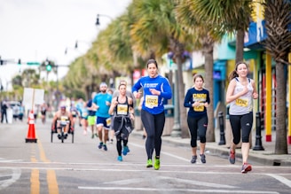 A group of runners, wearing numbered bibs, participate in a street race lined with palm trees. The scene is lively, with diverse participants including a person on a handcycle. The road is marked with orange cones, and spectators are visible in the background.