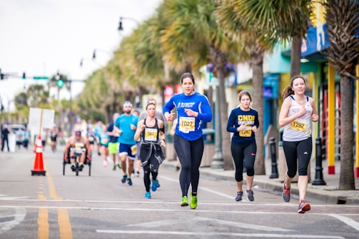 A group of runners, wearing numbered bibs, participate in a street race lined with palm trees. The scene is lively, with diverse participants including a person on a handcycle. The road is marked with orange cones, and spectators are visible in the background.