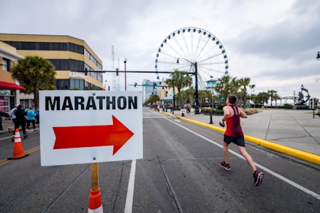A runner in a red tank top is racing down a city street with a large Ferris wheel in the background. A prominent sign with the word 'MARATHON' and an arrow points the direction, flanked by orange traffic cones. The street is lined with palm trees and buildings, suggesting an urban or coastal setting.