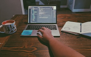 A person is working at a wooden desk with a laptop open to a code editor, showing lines of code. A hand is resting on the laptop's trackpad. Next to the laptop, there is an open notebook with writing and diagrams, and a pen resting on top. A cup filled with a beverage sits on the table, decorated with illustrations.