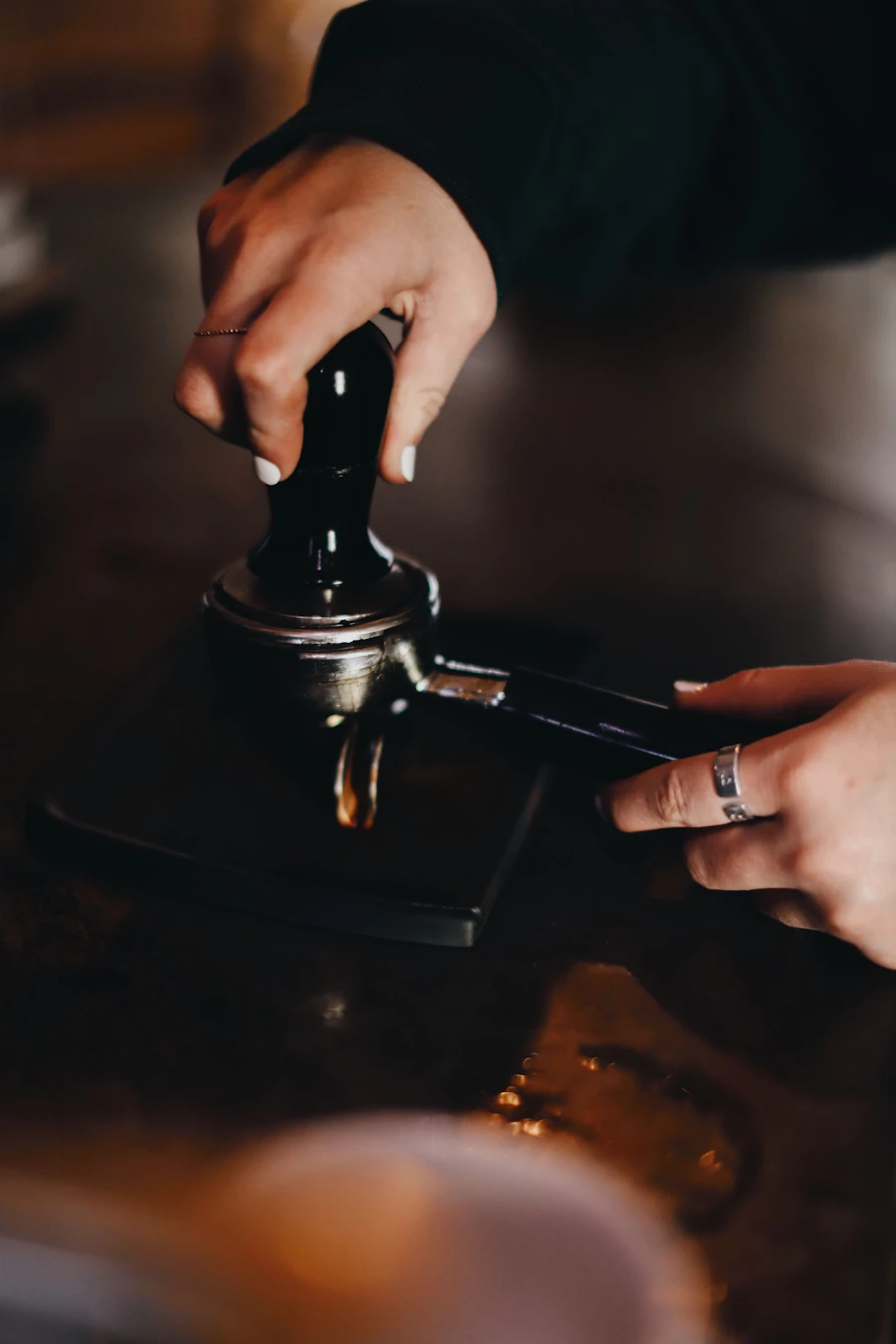Two hands levelling a tamper into a portafilter on a dark counter, one steadying the handle.