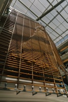 A large, vertical wooden structure is designed with a series of parallel slats. The pattern formed by the slats creates a wave-like texture, casting shadows on the surface. Natural light streams in from the glass ceiling, illuminating the installation.