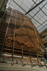 A large, vertical wooden structure is designed with a series of parallel slats. The pattern formed by the slats creates a wave-like texture, casting shadows on the surface. Natural light streams in from the glass ceiling, illuminating the installation.