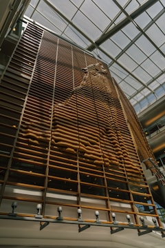 A large, vertical wooden structure is designed with a series of parallel slats. The pattern formed by the slats creates a wave-like texture, casting shadows on the surface. Natural light streams in from the glass ceiling, illuminating the installation.