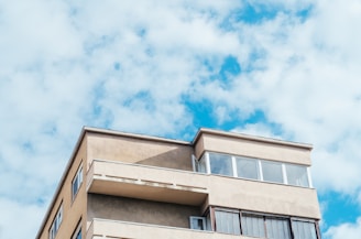 A modern apartment building with flat roofs and balconies is set against a background of a bright, partly cloudy sky. The structure appears to be of concrete with square windows and a minimalistic design.