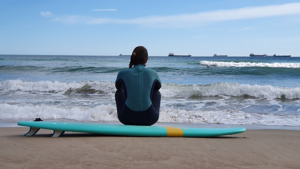 man in black wet suit sitting on blue surfboard on beach during daytime