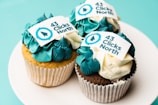 Three cupcakes with blue and white frosting are displayed on a white plate. Each cupcake features an edible decoration with the text '43 Clicks North', along with a logo in blue.