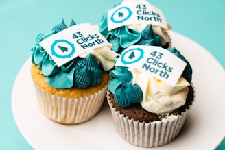 Three cupcakes with blue and white frosting are displayed on a white plate. Each cupcake features an edible decoration with the text '43 Clicks North', along with a logo in blue.