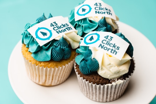 Three cupcakes with blue and white frosting are displayed on a white plate. Each cupcake features an edible decoration with the text '43 Clicks North', along with a logo in blue.