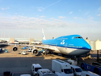 A large commercial airplane in KLM livery is parked at an airport gate. Several service vehicles and equipment can be seen around the aircraft on the tarmac. The sky is clear with some clouds, and the terminal is visible in the background.