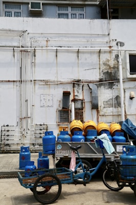 A somewhat rundown urban area features a collection of blue and yellow gas cylinders stacked on a bicycle cart. The background is an industrial-style building with visible pipes, air conditioning units, and a weathered paint job. The scene conveys a sense of practical, everyday urban life.