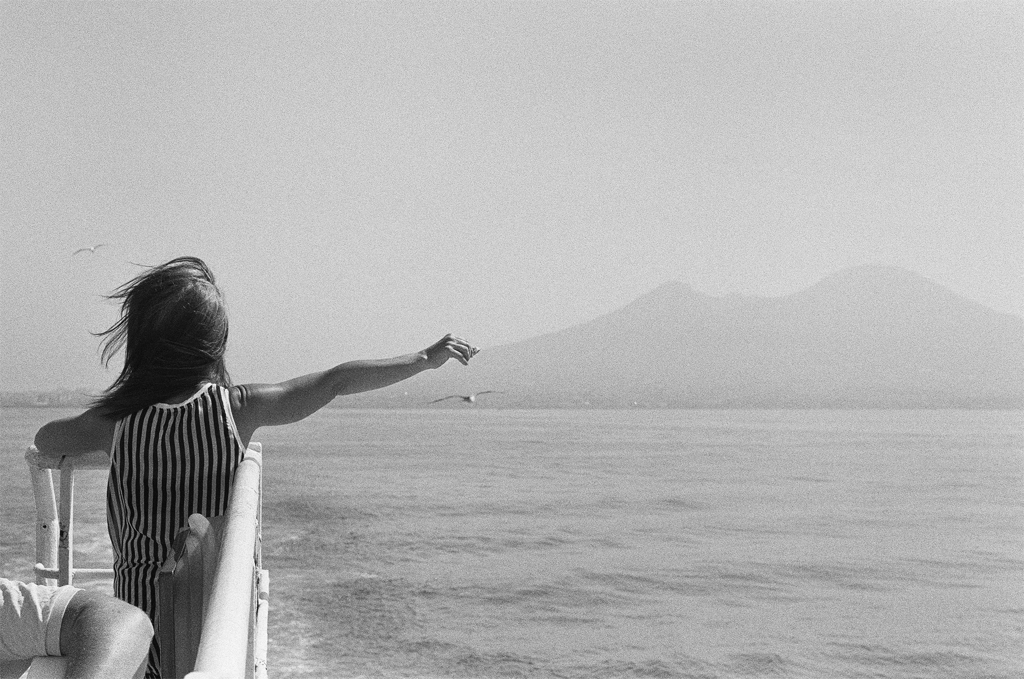 A woman in a boat is drawing the attention of seagulls that are following her. On the background, there is the Vesuvio, the volcano of Naples.