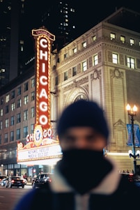 A brightly lit marquee with the word 'Chicago' in large letters extends vertically next to a classical-style building. The street is busy with cars, and a person in the foreground is out of focus, wearing a beanie and scarf.