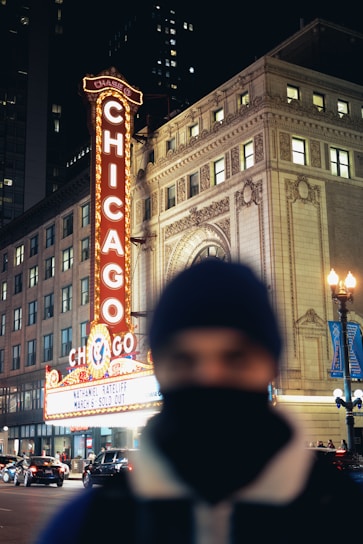 A brightly lit marquee with the word 'Chicago' in large letters extends vertically next to a classical-style building. The street is busy with cars, and a person in the foreground is out of focus, wearing a beanie and scarf.