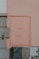 A wall featuring a rectangular area with wavy lines painted in a faded salmon color. The wall is divided into sections with different textures; a smoother peach-painted surface on the right and a rougher concrete section on the left. At the bottom, there are two utility boxes, one metallic and dark gray, the other lighter and more muted in color.