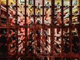 A well-organized wine rack filled with numerous bottles of wine, arranged horizontally and neatly labeled with letters. Various shades of red, brown, and amber are visible on the necks and bodies of the bottles. Soft lighting casts shadows and highlights the textures of the wooden rack.